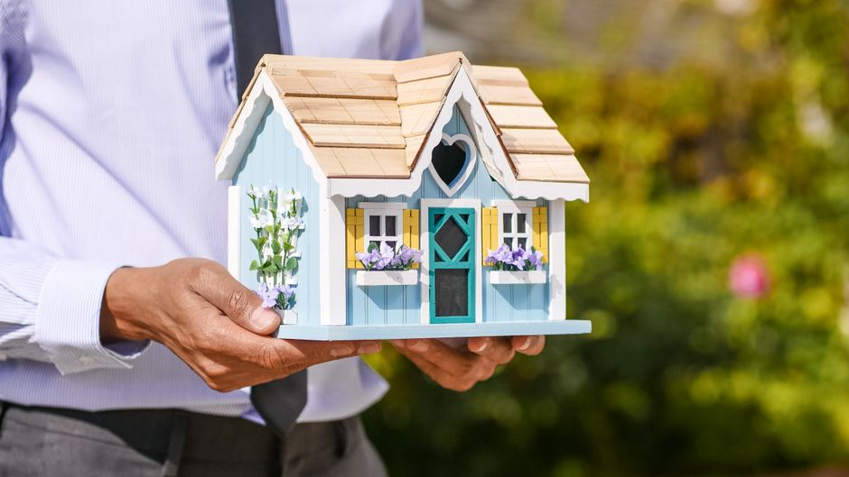 A person's hand carefully placing a small wooden house model onto a surface, illustrating the concept of building a real estate portfolio or making a new investment.