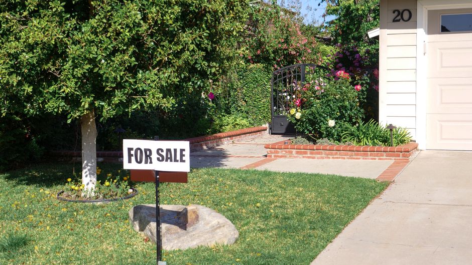 A homeowner puts in the work, hammering a 'For Sale' sign into the green lawn in front of their suburban house, illustrating the do-it-yourself nature of an FSBO listing.