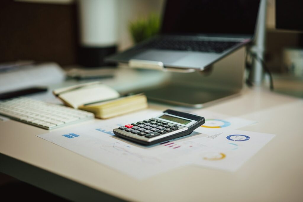 Person sits at a bright, minimalist desk, thoughtfully using a calculator next to a laptop and house keys, symbolizing the...