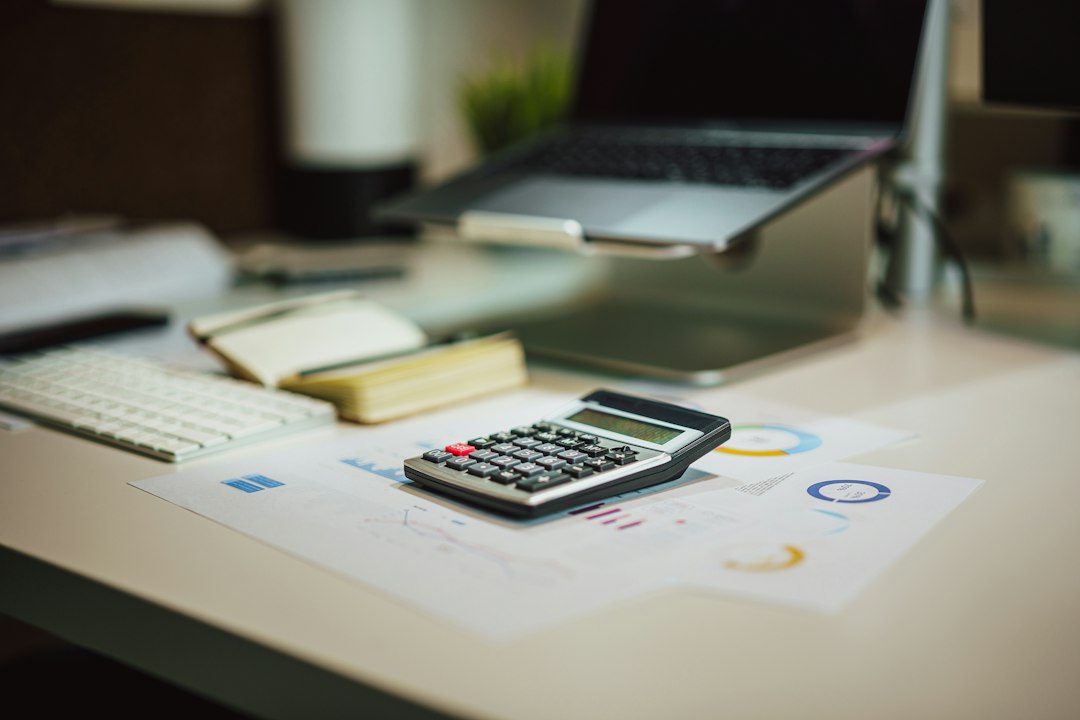 Person sits at a bright, minimalist desk, thoughtfully using a calculator next to a laptop and house keys, symbolizing the...